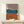 May include: Abstract geometric art print in wooden frame showing three overlapping organic shapes in taupe, teal, and terracotta orange against cream background. Artwork displayed on light wood shelf with small white wrapped gift decorated with evergreen sprig. Soft natural lighting on neutral wall.