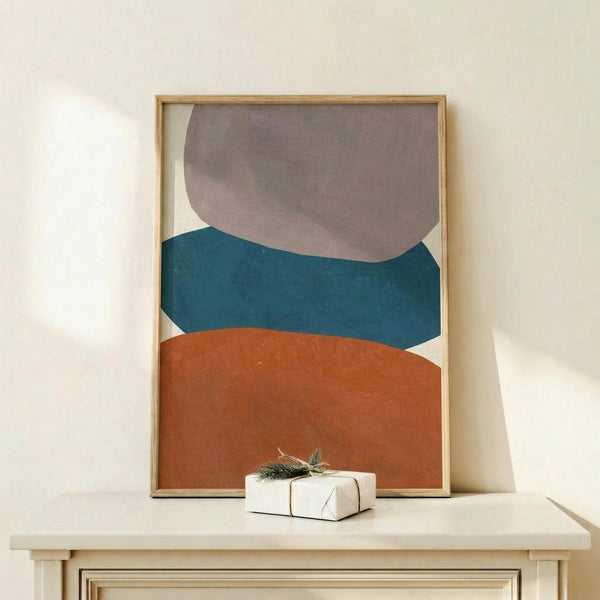 May include: Abstract geometric art print in wooden frame showing three overlapping organic shapes in taupe, teal, and terracotta orange against cream background. Artwork displayed on light wood shelf with small white wrapped gift decorated with evergreen sprig. Soft natural lighting on neutral wall.
