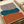 Abstract geometric artwork featuring three organic, curved shapes in stacked layers - taupe gray at top, teal blue in middle, and burnt orange at bottom - painted on cream background. The artwork sits flat on a light wooden surface next to rolled paper tubes, showing the poster's actual size and matte finish texture.