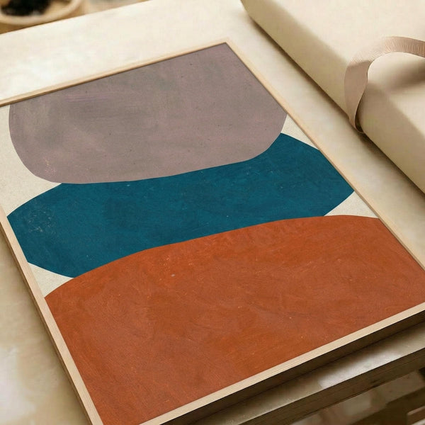 Abstract geometric artwork featuring three organic, curved shapes in stacked layers - taupe gray at top, teal blue in middle, and burnt orange at bottom - painted on cream background. The artwork sits flat on a light wooden surface next to rolled paper tubes, showing the poster's actual size and matte finish texture.