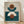 May include: Framed geometric abstract art print leaning against white textured wall on light wood floor. The artwork features layered semi-circles and circles in terracotta brown, cream, and teal blue tones on a neutral beige background, displayed in a natural wood frame. Soft natural lighting creates gentle shadows across the scene.
