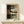 May include: Abstract art print in light wood frame displayed on light wooden surface against cream wall. Artwork features geometric shapes in earth tones - blacks, browns, tans, and grays - with curved black lines creating contemporary composition. Small white wrapped gift with pine sprig sits beside the framed piece. Soft natural lighting creates gentle shadows on the minimalist scene.