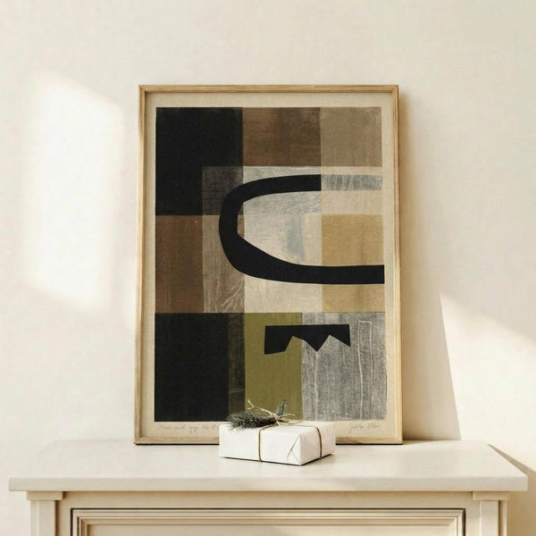 May include: Abstract art print in light wood frame displayed on light wooden surface against cream wall. Artwork features geometric shapes in earth tones - blacks, browns, tans, and grays - with curved black lines creating contemporary composition. Small white wrapped gift with pine sprig sits beside the framed piece. Soft natural lighting creates gentle shadows on the minimalist scene.