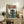 May include: Framed anime-style poster leaning against white wall on rustic wooden sideboard, showing figure in brown coat standing before large green mech robot in futuristic cityscape. Japanese text at top in orange and black. Room has beige armchair, fern plant, ceramic vase, and stacked books creating cozy living space setting.
