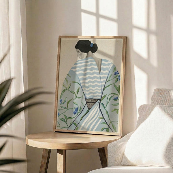 May include: Framed artwork displayed on a round wooden side table next to a beige upholstered chair and plant. The illustration shows a woman from behind wearing a light blue and white striped garment with floral botanical details featuring green leaves and small blue flowers. She has dark hair styled in a low bun. The piece has a soft, watercolor-like quality with pale blue and green tones against a cream background, presented in a thin gold frame.