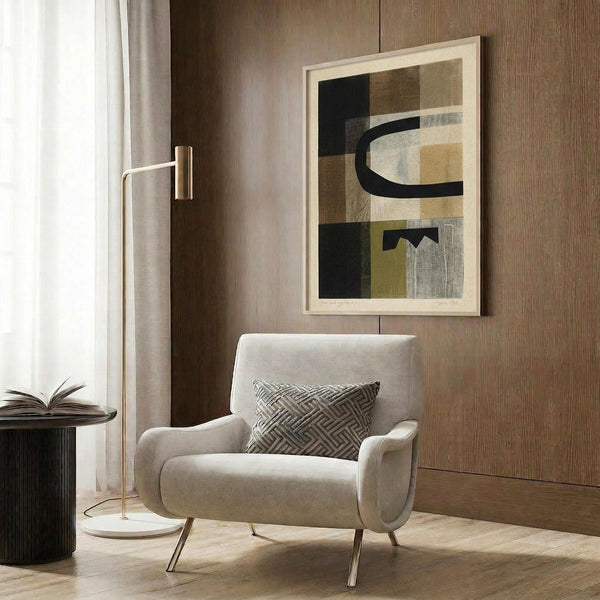 May include: Abstract geometric artwork in light wood frame hanging on wood paneled wall above cream upholstered armchair. Print features overlapping rectangular shapes in muted tones of black, beige, olive green, and gray with curved black lines. Modern reading corner with brass floor lamp, patterned throw pillow, and dark round side table with open book.