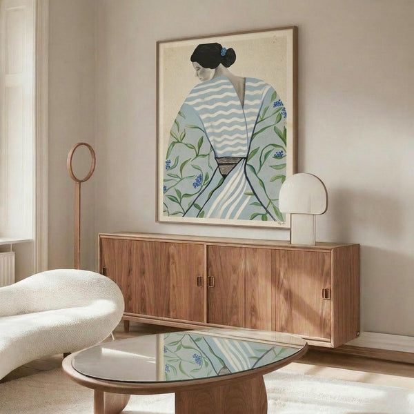 May include: Framed artwork showing a figure in traditional Asian dress with black hair, wearing a striped blue and white garment with floral outer robe featuring green leaves and small blue flowers. The print hangs above a mid-century modern walnut credenza with sliding doors. Scene includes a white curved chair, brass floor lamp, modern table lamp, and oval glass coffee table reflecting the artwork, all in a bright room with white walls.