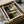 Abstract geometric artwork with organic curved shapes in earth tones of brown, beige, olive green, and black, displayed in a black frame within an open cardboard shipping box. The print is surrounded by protective tissue paper and wooden dowels, showing how the artwork arrives packaged for shipping.