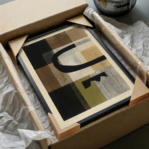Abstract geometric artwork with organic curved shapes in earth tones of brown, beige, olive green, and black, displayed in a black frame within an open cardboard shipping box. The print is surrounded by protective tissue paper and wooden dowels, showing how the artwork arrives packaged for shipping.