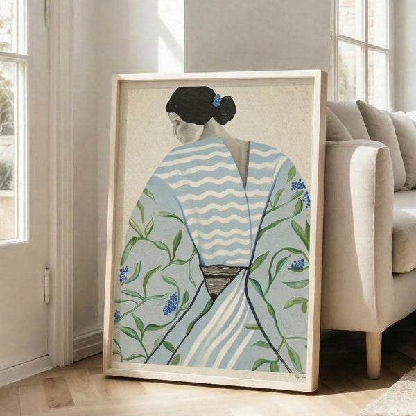 May include: Large framed artwork leaning against white wall beside beige sofa in bright living room with windows. The poster shows a woman in profile with black upswept hair and blue flower accent, wearing a light blue and white striped robe decorated with green botanical branches and small blue berries. The illustration has a soft, minimalist style with muted colors on cream background in natural wood frame.