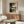 May include: Abstract geometric poster with earthy tones including black, brown, olive green, and cream blocks with curved black line, displayed in thin wooden frame above mid-century walnut credenza. Modern living room setting features white textured chair, oval glass coffee table, and minimalist table lamp on neutral walls.