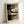 Framed abstract art print featuring geometric shapes in muted earth tones including black, brown, beige, olive green, and gray. The composition shows overlapping rectangular forms with curved black lines creating organic flowing elements against angular backgrounds. The artwork is displayed in a light wood frame, leaning against a white wall on what appears to be a white surface. The print has a contemporary minimalist style with subtle textures and layered translucent effects throughout the geo