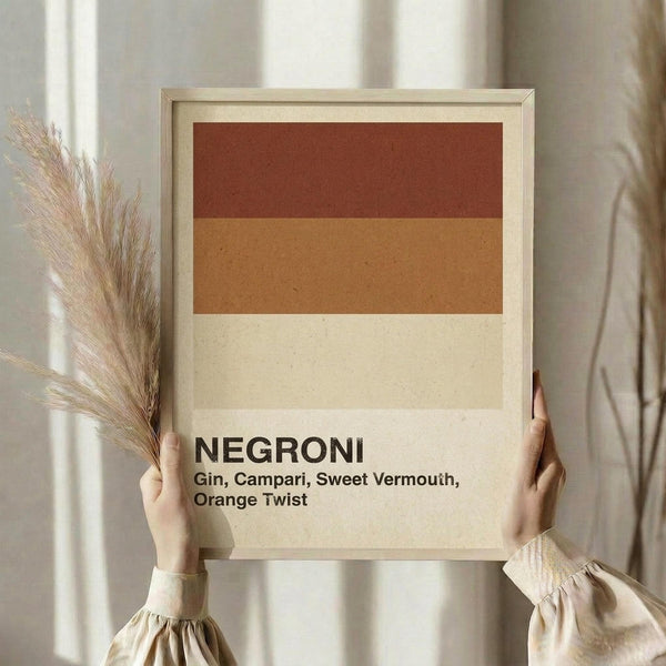 May include: Hands holding a framed minimalist poster with horizontal color blocks in rust red, warm orange, and cream. Bold black text reads "NEGRONI" with cocktail ingredients listed below. Light wood frame, neutral beige setting with dried pampas grass visible to the left.