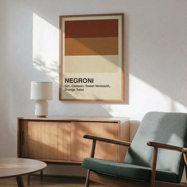 May include: Framed poster with horizontal color bands in burgundy, tan, and cream showing "NEGRONI" text and cocktail ingredients, displayed above mid-century modern wooden credenza with table lamp, next to sage green upholstered armchair with wood frame in sunlit room with white walls