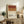 May include: Framed poster displaying color-blocked bands in burgundy, orange, and cream with "NEGRONI" and cocktail ingredients text below. Wooden frame sits on rustic wood sideboard alongside books, pottery vase, and fern plant. Neutral living room setting with beige armchair visible.