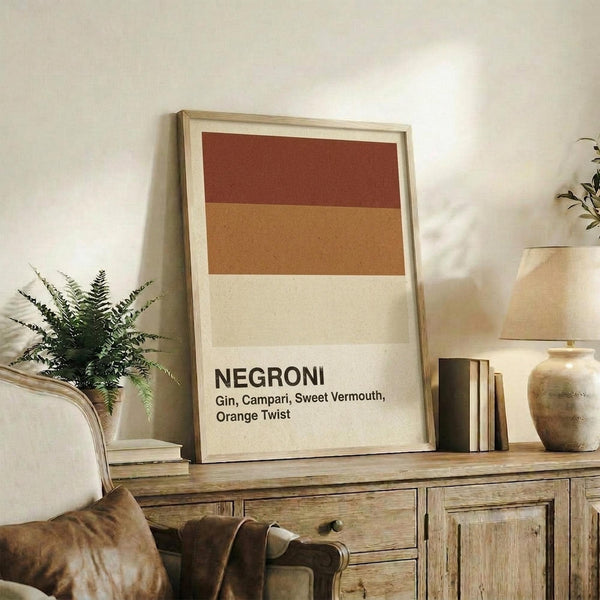 May include: Framed poster displaying color-blocked bands in burgundy, orange, and cream with "NEGRONI" and cocktail ingredients text below. Wooden frame sits on rustic wood sideboard alongside books, pottery vase, and fern plant. Neutral living room setting with beige armchair visible.