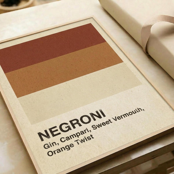 Minimalist poster print featuring horizontal color stripes in rust red, warm orange, and cream representing Negroni cocktail ingredients. Bold black text below reads "NEGRONI" with smaller text listing "Gin, Campari, Sweet Vermouth, Orange Twist". The print has a clean modern design with earthy warm tones on cream background, shown flat against neutral wrapping paper with beige ribbon visible at edge.