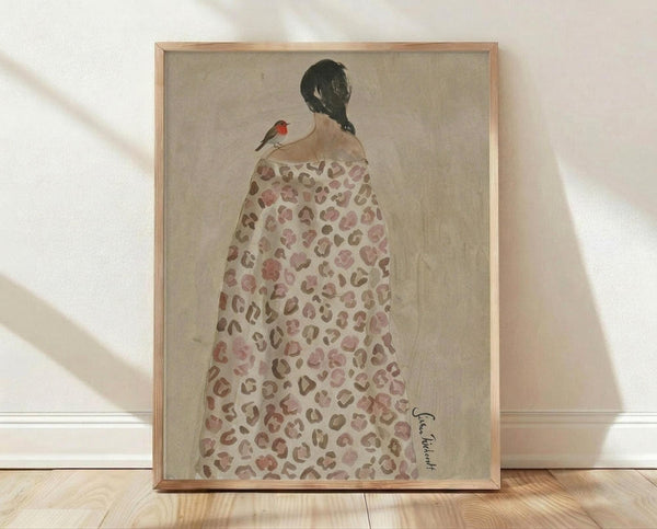May include: Framed art print leaning against white wall on light wood floor with natural lighting. Shows woman from behind wearing leopard print dress or robe with dark braided hair and small red bird perched on her shoulder. Artwork has muted earth tones on beige background in thin natural wood frame.