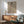May include: Framed artwork showing a woman in leopard print robe with small red bird on her shoulder, displayed above wooden sideboard with white ceramic lamp in modern living room setting with sage green armchair and natural lighting creating soft shadows on white walls.