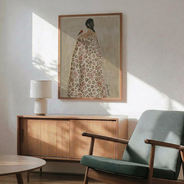 May include: Framed artwork showing a woman in leopard print robe with small red bird on her shoulder, displayed above wooden sideboard with white ceramic lamp in modern living room setting with sage green armchair and natural lighting creating soft shadows on white walls.