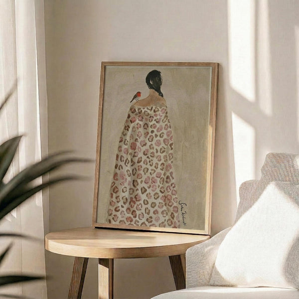 May include: Framed art print displayed on round wooden side table next to beige upholstered chair. Artwork shows woman from behind wearing leopard print blanket or robe with small red bird perched on her shoulder, painted in soft earth tones against cream background. Natural lighting creates shadows across neutral-toned room with plant visible in corner.