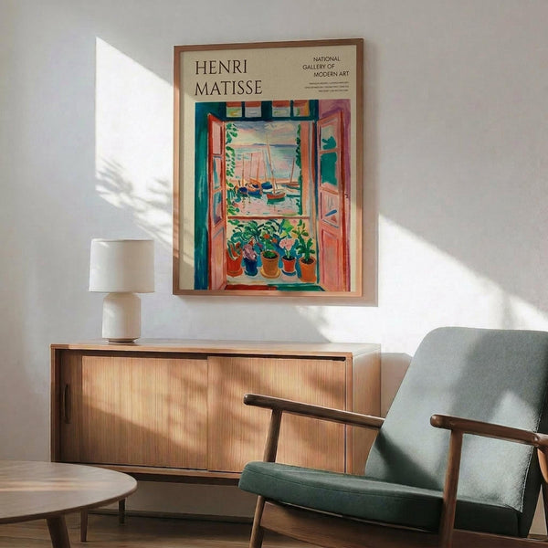 May include: Henri Matisse exhibition poster showing colorful Fauve-style painting of open window with harbor view and potted plants on windowsill. Cream background with black text reading "HENRI MATISSE" and "NATIONAL GALLERY OF MODERN ART". Displayed in thin wooden frame above mid-century modern walnut credenza with white ceramic lamp. Sage green upholstered armchair with wooden frame sits nearby. Warm sunlight creates dramatic shadows on white wall.