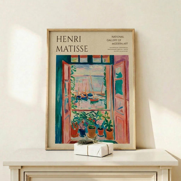 May include: Framed Henri Matisse exhibition poster featuring colorful Fauve-style painting of open window with boats visible outside and potted plants on windowsill. Poster displayed on light wood shelf with small wrapped gift box beside it. Bright blues, oranges, greens and pinks dominate the artwork within thin wooden frame against cream wall.