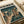 May include: Vintage-style poster print displayed on white bedding showing anime character in tan coat standing on futuristic city rooftop at night. Dark teal and orange color palette with Japanese text header. Rain falls over neon-lit skyscrapers. Poster appears aged with cream borders, lying flat against white fabric background.