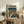 May include: Framed anime-style poster featuring a character in a brown coat standing on a futuristic city street with tall buildings and glowing signs, displayed on a rustic wooden console table alongside books, a ceramic vase, and fern plant in a cozy living room setting with beige armchair