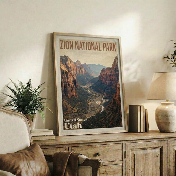 May include: Vintage-style travel poster featuring Zion National Park's dramatic red rock canyon landscape with winding river below, displayed in light wood frame on rustic wooden sideboard alongside fern plant, books, ceramic vase and table lamp in warm neutral living room setting