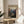 May include: Framed vintage-style travel poster featuring a dramatic canyon landscape with red rock cliffs, winding river, and mountains under golden lighting. The poster sits on a round wooden side table next to a beige upholstered chair, with natural sunlight streaming across white walls and a potted plant visible in the corner of this modern living space.