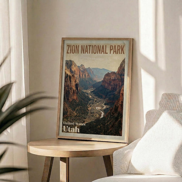 May include: Framed vintage-style travel poster featuring a dramatic canyon landscape with red rock cliffs, winding river, and mountains under golden lighting. The poster sits on a round wooden side table next to a beige upholstered chair, with natural sunlight streaming across white walls and a potted plant visible in the corner of this modern living space.