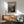 May include: Framed vintage-style travel poster featuring dramatic canyon landscape with winding river, red rock formations, and mountain vista. Warm earth tones complement mid-century modern living room with sage green armchair, walnut sideboard, white ceramic lamp, and natural lighting creating geometric shadows on white walls.