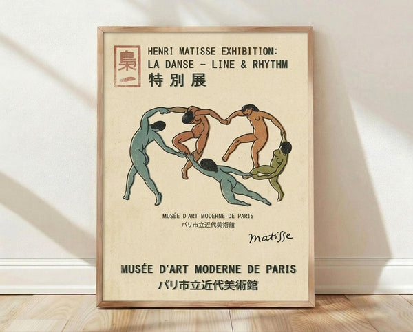 May include: A framed vintage exhibition poster featuring five stylized nude dancers in a circular formation rendered in muted tones of green, brown, and beige. The poster displays text in English, French and Japanese for a Henri Matisse exhibition at Musée d'Art Moderne de Paris. The artwork is shown in a light wood frame, leaning against a white wall on wooden flooring with soft natural lighting creating subtle shadows.