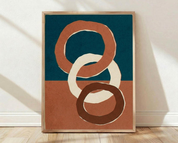 May include: Framed abstract art print featuring three interlocking oval rings in terracotta orange, cream, and dark brown against a split background of teal blue and dusty rose. The wooden frame sits on light hardwood flooring against white fabric backdrop with soft natural lighting creating gentle shadows.
