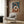 May include: Modern beige upholstered chair with geometric patterned pillow positioned in front of wood-paneled wall. Abstract art print featuring interlocking circular rings in terracotta orange, teal blue, cream and brown colors within light wood frame. Gold floor lamp and dark side table with open book visible. Warm neutral interior with natural lighting from window.