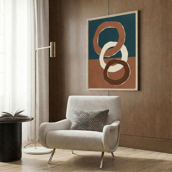 May include: Modern beige upholstered chair with geometric patterned pillow positioned in front of wood-paneled wall. Abstract art print featuring interlocking circular rings in terracotta orange, teal blue, cream and brown colors within light wood frame. Gold floor lamp and dark side table with open book visible. Warm neutral interior with natural lighting from window.