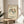 May include: Framed vintage-style Matisse exhibition poster featuring stylized dancing figures in green, brown, and orange tones against cream background, displayed on round wooden stool beside beige upholstered chair with white pillow, plant visible in corner, soft natural lighting creating gentle shadows on neutral walls.