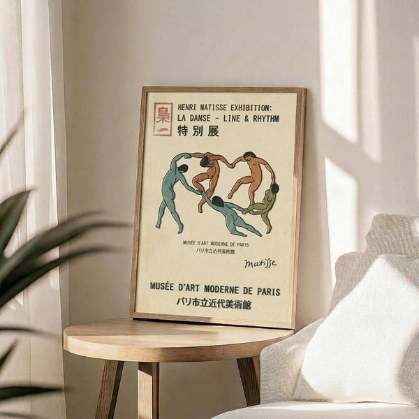 May include: Framed vintage-style Matisse exhibition poster featuring stylized dancing figures in green, brown, and orange tones against cream background, displayed on round wooden stool beside beige upholstered chair with white pillow, plant visible in corner, soft natural lighting creating gentle shadows on neutral walls.