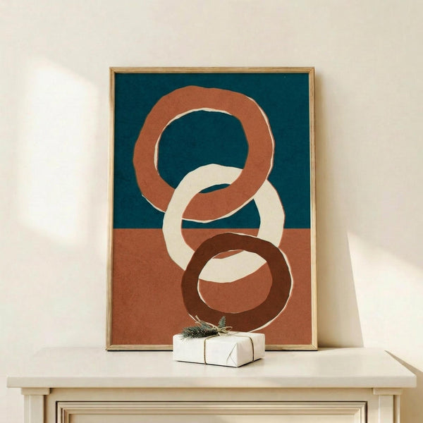 May include: Modern abstract art print in wooden frame displaying three interlocking rings in terracotta, cream, and dark brown colors on split teal and rust background, displayed on light wooden console table beside small white wrapped gift with evergreen sprig against cream wall