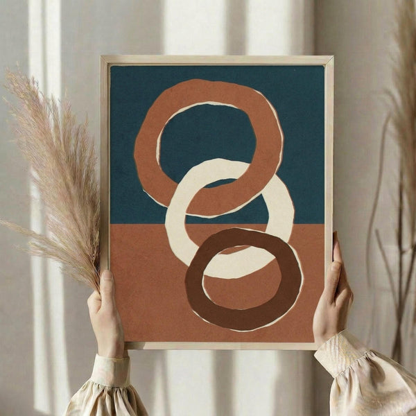 May include: Person in cream sweater holding framed abstract art print featuring three interlocking oval rings in terracotta, cream, and dark brown against split teal and terracotta background. One hand also holds dried pampas grass stems. Natural lighting from window creates soft shadows on white curtained wall.