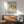 May include: Framed Matisse exhibition poster featuring dancing figures in green and orange tones, displayed above a wooden sideboard with white ceramic lamp. Mid-century modern living room with teal upholstered armchair, round wood coffee table, and natural sunlight creating shadows on white wall.