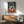 May include: Abstract geometric poster with interlocking circular rings in terracotta, cream, and dark brown colors on teal and beige split background, displayed in thin wood frame above mid-century modern credenza. Room features sage green upholstered armchair with wooden arms, white ceramic table lamp, and warm natural lighting creating soft shadows on white wall.