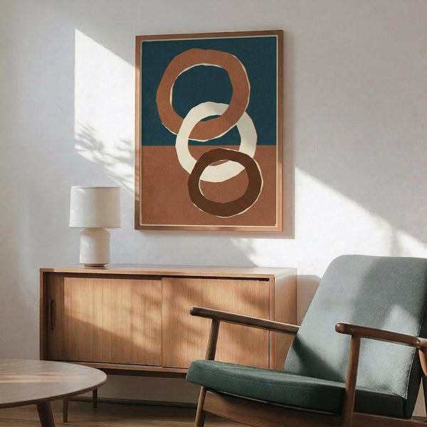 May include: Abstract geometric poster with interlocking circular rings in terracotta, cream, and dark brown colors on teal and beige split background, displayed in thin wood frame above mid-century modern credenza. Room features sage green upholstered armchair with wooden arms, white ceramic table lamp, and warm natural lighting creating soft shadows on white wall.