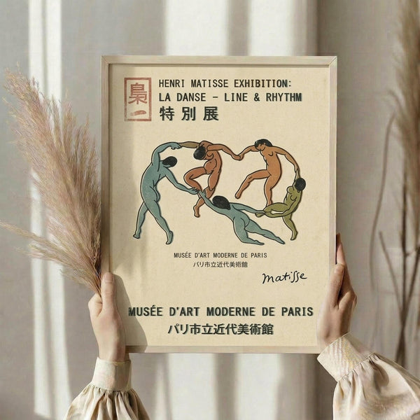 May include: Person in beige ruffled sleeves holding framed exhibition poster featuring dancing figures in green, brown and cream tones. Light wood frame against neutral curtained background. Left hand holds dried pampas grass stems. Poster shows circular arrangement of stylized nude dancers with French and Japanese museum text.