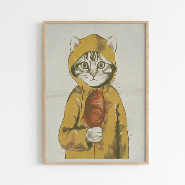 Whimsical illustration of a tabby cat wearing a bright yellow hooded raincoat, holding a red fish in its paws. The cat has large green eyes and its ears peek through holes in the hood. The artwork is printed on textured cream paper with a weathered, vintage appearance and displayed in a light wood frame against a white wall.