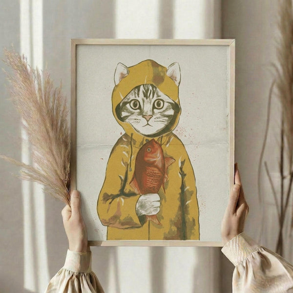 May include: Person holding framed artwork showing a tabby cat wearing a yellow hooded raincoat and holding a red fish, displayed in a light wood frame against cream textured paper background. Dried pampas grass visible on left side, soft natural lighting from window creates gentle shadows on white curtains and neutral room setting.