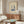 May include: Framed beige poster with blue illustrated figures dancing on a turntable, displayed above a walnut mid-century credenza with brass hardware. Modern white curved chair and round glass coffee table with colorful abstract art visible. White mushroom table lamp and brass floor lamp complete the minimalist living room scene with natural lighting.