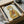 May include: Framed poster print in black frame showing a tabby cat wearing a yellow hooded raincoat and holding a red fish. The artwork has a vintage, illustrated style on cream-colored paper. Frame is packaged in brown cardboard box with white tissue paper protection, sitting on white fabric surface.