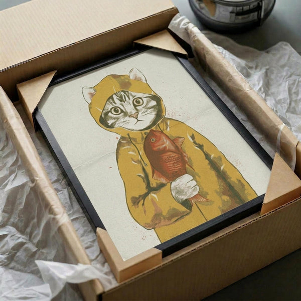 May include: Framed poster print in black frame showing a tabby cat wearing a yellow hooded raincoat and holding a red fish. The artwork has a vintage, illustrated style on cream-colored paper. Frame is packaged in brown cardboard box with white tissue paper protection, sitting on white fabric surface.